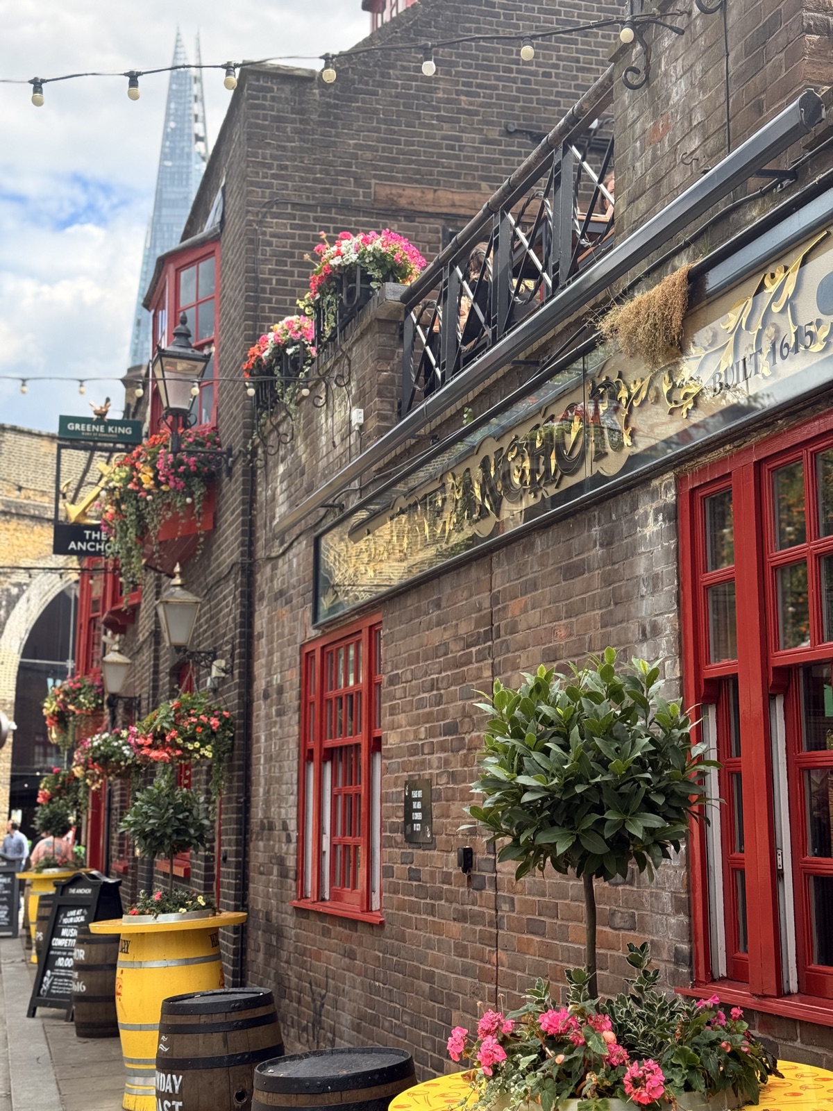 The Anchor pub in Southwark with brick walls, red window frames, hanging flower baskets, and the Shard rising behind