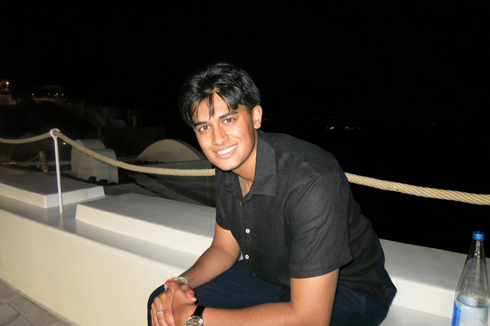 Ariyan Patel smiling at night in Santorini, white rooftops and a rope railing behind him