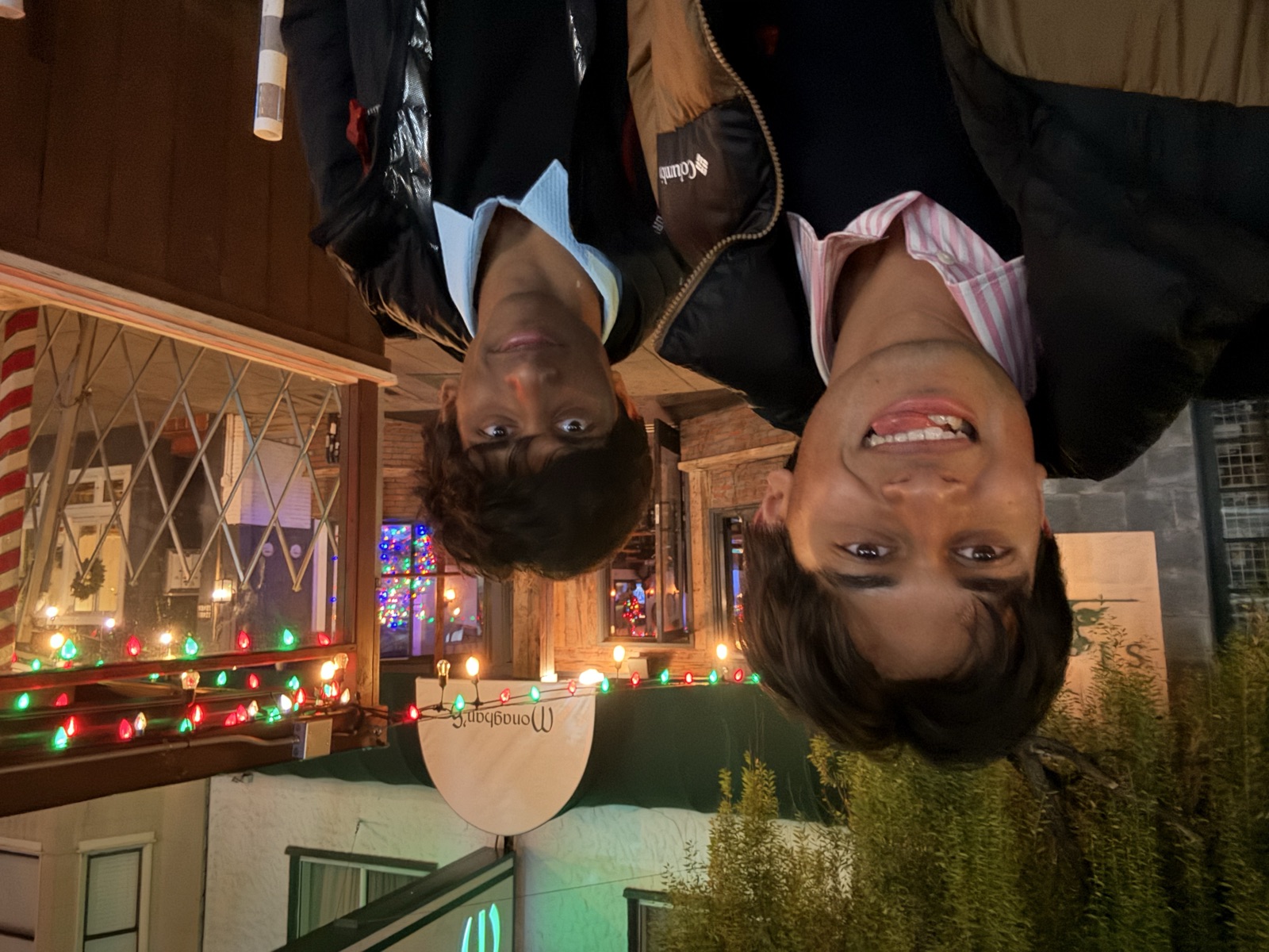 Ariyan and Kush grinning at night outside a bar strung with red and green holiday lights